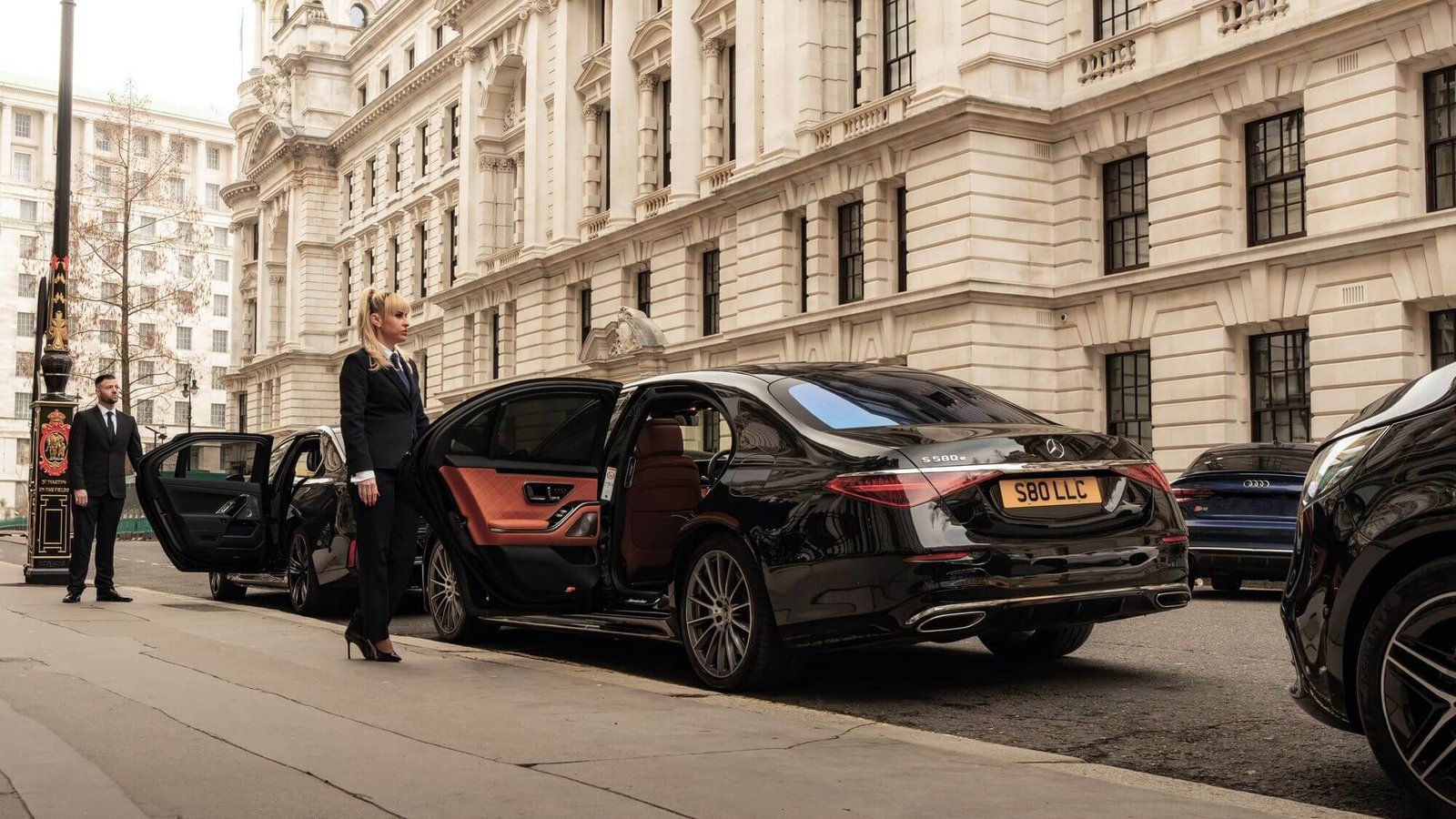 Luxury chauffeur in a suit holding open the car door for a customer exiting a luxury vehicle in central London's city centre.